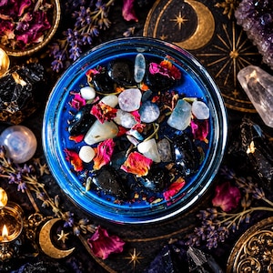Direct overhead view of the Hecate ritual-inspired candle featuring saturated cobalt blue wax, black obsidian crystals, pale moonstone pieces, dried pink and red roses, and botanical herbs. The glass jar is arranged against a dark mystical backdrop decorated with antique gold crescent moons, celestial starbursts, purple lavender, crystals, and burgundy flowers. An ornate handmade goddess candle celebrating Hecate, crossroads magic, the dark moon, personal transformation, and spiritual protection