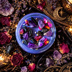 A round blue candle in a glass jar topped with purple amethyst crystals, black obsidian stones, and dried pink rose petals. The candle wax is a deep blue hue, and the surface is decorated with these natural elements.