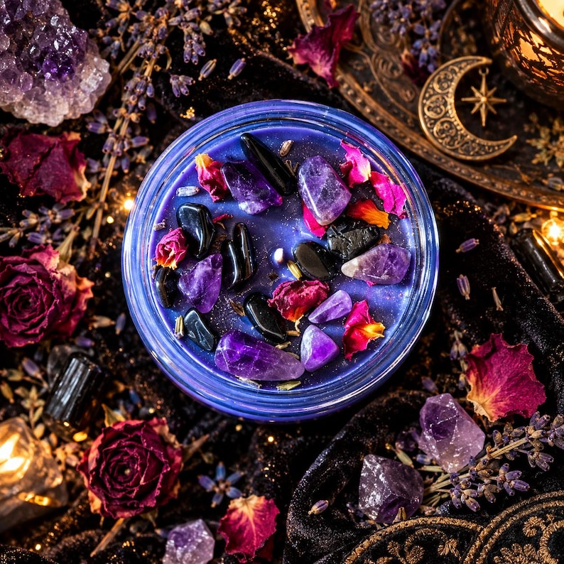 A round blue candle in a glass jar topped with purple amethyst crystals, black obsidian stones, and dried pink rose petals. The candle wax is a deep blue hue, and the surface is decorated with these natural elements.