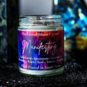 Op de afbeelding: Een glazen potkaars met een kleurrijk etiket. Het etiket luidt "Enchanted Moon Crystal Manifesting" met een lijst van ingredi&euml;nten. De kaars is met de hand gegoten in Texas. De achtergrond is donker en wazig.