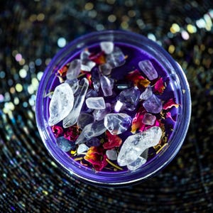 Pu&ograve; includere: Una candela viola in un contenitore di vetro trasparente, adornata con cristalli trasparenti e viola, petali di rosa secchi ed erbe. La candela ha una superficie liscia ed &egrave; circondata da uno sfondo scuro e scintillante.
