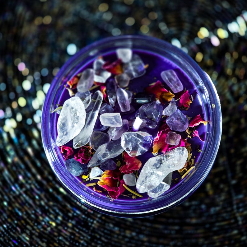 Pu&ograve; includere: Una candela viola in un contenitore di vetro trasparente, adornata con cristalli trasparenti e viola, petali di rosa secchi ed erbe. La candela ha una superficie liscia ed &egrave; circondata da uno sfondo scuro e scintillante.