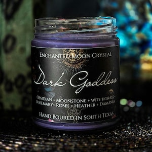 May include: A clear glass jar candle filled with deep purple wax. The black label features the text Enchanted Moon Crystal, Dark Goddess, and a list of ingredients including Obsidian, Moonstone, Witchgrass, Rosemary, Roses, Heather, and Damania.
