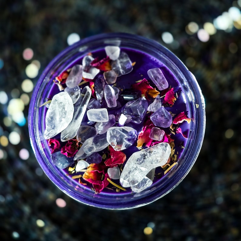 Pu&ograve; includere: Una candela viola in un barattolo di vetro, riempita con cristalli trasparenti e ametista, e petali di rosa secchi. La candela &egrave; vista dall'alto, con uno sfondo scuro e macchiettato.