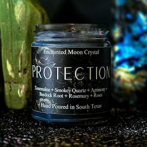 May include: A glass jar candle with a black label featuring the text Enchanted Moon Crystal, PROTECTION, and a list of ingredients including Tourmaline, Smokey Quartz, Agrimony, Burdock Root, Rosemary, and Roses, with Hand Poured in South Texas.