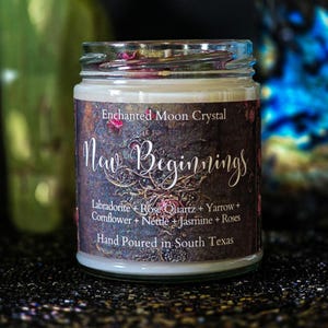 May include: A glass jar candle with a label that reads "Enchanted Moon Crystal New Beginnings." The label lists ingredients like Labradorite, Rose Quartz, and Jasmine. The candle is hand-poured in South Texas.