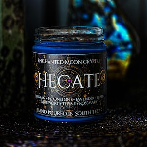 Handmade Hecate intention candle in a clear glass jar with rich blue wax and an ornate black and gold celestial label. Moonstone, Lavender, Mugwort, Thyme, and Rosemary decorate the candle for a dark, mystical botanical appearance.