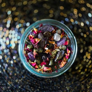 Detailed overhead photograph of the Someone F'd Up candle showing dried flowers, rose petals, herbs, crystal pieces, and stones layered over the wax. Keywords: dressed candle, herbs and crystals, spellwork, ritual candle, witchcraft.