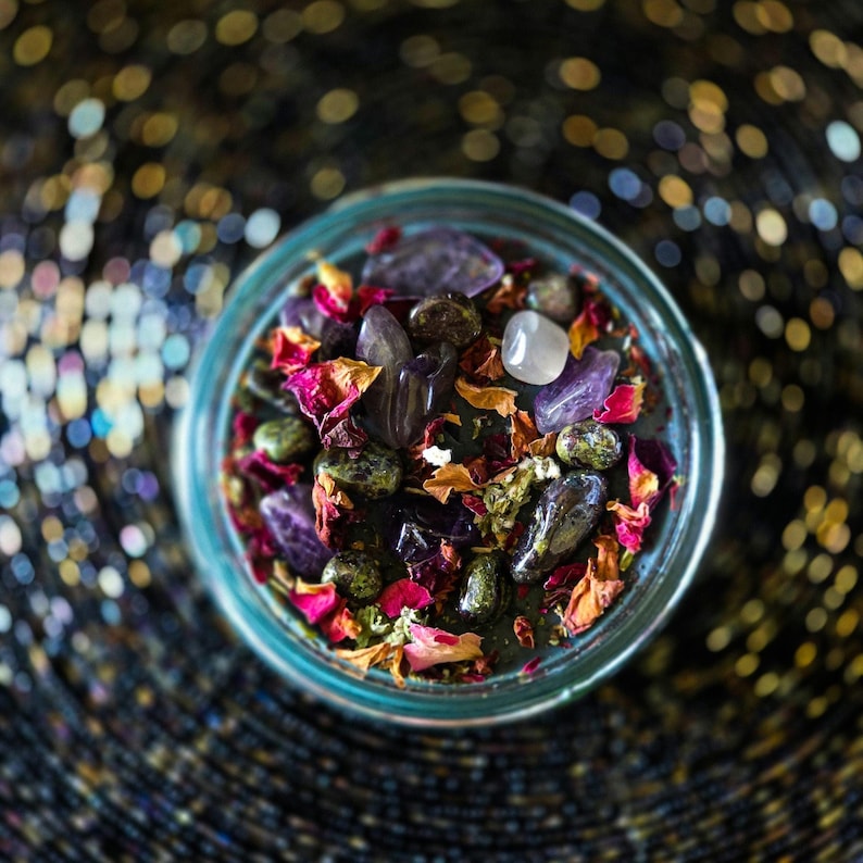 Detailed overhead photograph of the Someone F'd Up candle showing dried flowers, rose petals, herbs, crystal pieces, and stones layered over the wax. Keywords: dressed candle, herbs and crystals, spellwork, ritual candle, witchcraft.