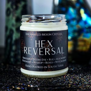 May include: A clear glass candle jar filled with white wax, featuring a black label with the text Enchanted Moon Crystal, Hex Reversal, and a list of ingredients including Obsidian, Tigers Eye, Rue, Mugwort, Rosemary, Hyssop, Roses, and Witchgrass.
