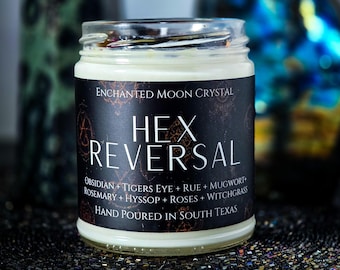 May include: A clear glass candle jar filled with white wax, featuring a black label with the text Enchanted Moon Crystal, Hex Reversal, and a list of ingredients including Obsidian, Tigers Eye, Rue, Mugwort, Rosemary, Hyssop, Roses, and Witchgrass.