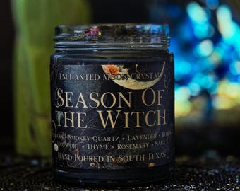 May include: A black glass candle jar with a dark label featuring gold text that reads Enchanted Moon Crystal Season of the Witch. The label lists ingredients including smokey quartz, lavender, roses, mugwort, thyme, rosemary, and sage.