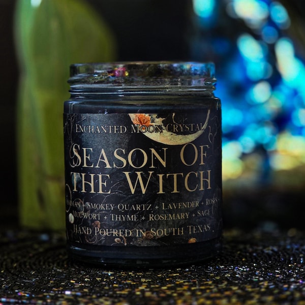 May include: A black glass candle jar with a dark label featuring gold text that reads Enchanted Moon Crystal Season of the Witch. The label lists ingredients including smokey quartz, lavender, roses, mugwort, thyme, rosemary, and sage.