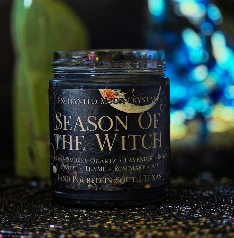 Season of the Witch candle photographed against a dark mystical background with luminous blue and green accents. Black wax and a vintage botanical label create a moody witchy aesthetic for this handmade ritual and intention candle.