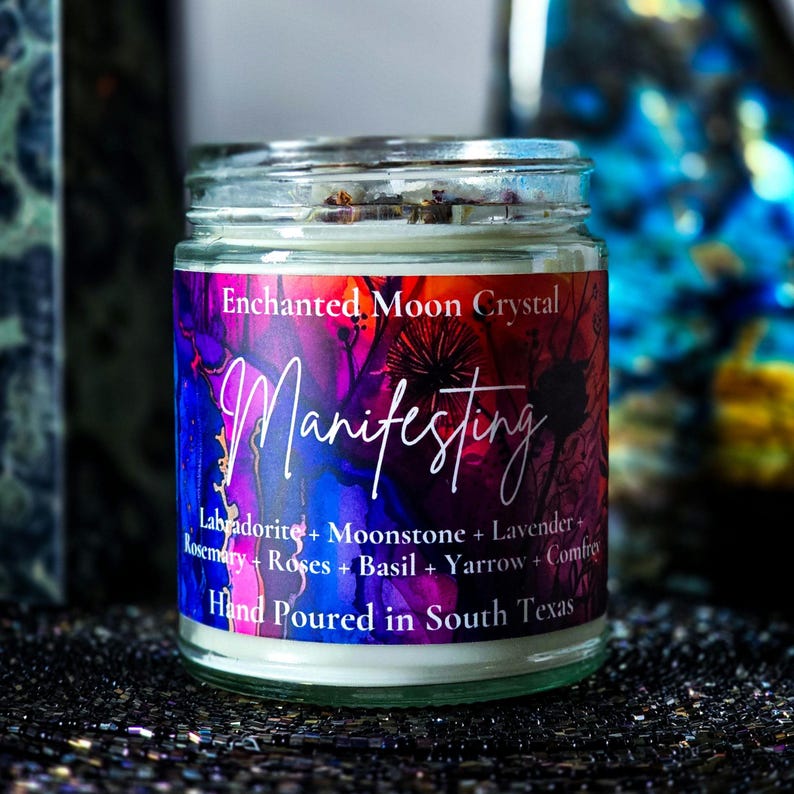Op de afbeelding: Een glazen potkaars met een kleurrijk etiket met de tekst "Enchanted Moon Crystal Manifesting". Het etiket bevat ingredi&euml;nten zoals Labradoriet, Maansteen en Lavendel. De kaars is met de hand gegoten in South Texas.