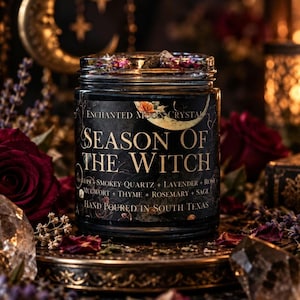 Season of the Witch hand-poured candle Enchanted Moon Crystal celestial label and botanical toppings visible above the glass rim. Surrounded by dark red roses, smoky quartz crystals, purple lavender, ornate brass accents, golden crescent moon decorations, and warm candlelight. Moody gothic candle photography highlighting a handcrafted spiritual intention candle inspired by protection, grounding, witchcraft, autumn rituals, and magical home decor.
