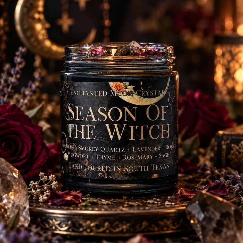 Season of the Witch hand-poured candle Enchanted Moon Crystal celestial label and botanical toppings visible above the glass rim. Surrounded by dark red roses, smoky quartz crystals, purple lavender, ornate brass accents, golden crescent moon decorations, and warm candlelight. Moody gothic candle photography highlighting a handcrafted spiritual intention candle inspired by protection, grounding, witchcraft, autumn rituals, and magical home decor.