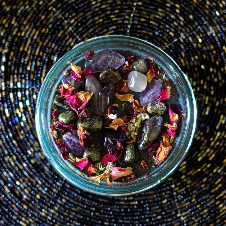 Close overhead view of the Someone F'd Up candle featuring dried rose petals, botanical herbs, crystal chips, and decorative stones arranged across the dark wax. Keywords: dressed candle, ritual herbs, crystals, spell candle, witchcraft.