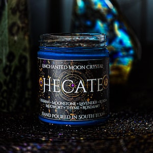 May include: A blue candle in a glass jar with a black label featuring the text ENCHANTED MOON CRYSTAL HECATE, along with a list of ingredients including obsidian, moonstone, lavender, roses, mugwort, thyme, and rosemary, and the text HAND POURED IN SOUTH TEXAS.