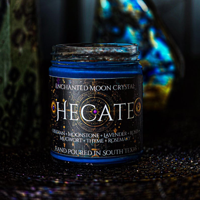 May include: A blue candle in a glass jar with a black label featuring the text ENCHANTED MOON CRYSTAL HECATE, along with a list of ingredients including obsidian, moonstone, lavender, roses, mugwort, thyme, and rosemary, and the text HAND POURED IN SOUTH TEXAS.