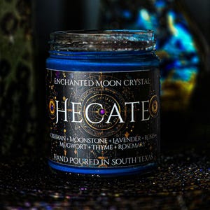 Hand-poured Hecate candle featuring rich blue wax in a clear glass jar with a black celestial label. Topped with Moonstone, Lavender, Mugwort, Thyme, and Rosemary for a colorful mystical finish. Keywords: goddess candle, Hecate, spiritual candle.