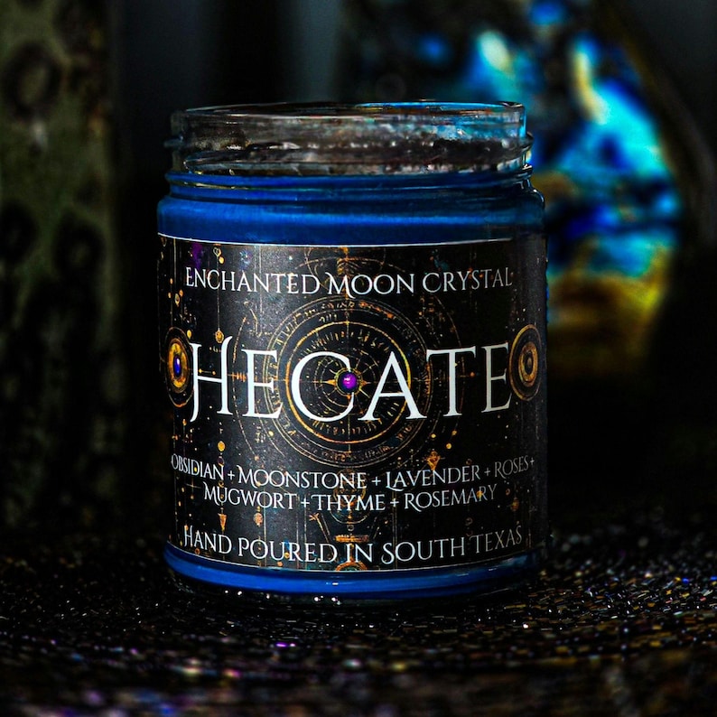 Hand-poured Hecate candle featuring rich blue wax in a clear glass jar with a black celestial label. Topped with Moonstone, Lavender, Mugwort, Thyme, and Rosemary for a colorful mystical finish. Keywords: goddess candle, Hecate, spiritual candle.