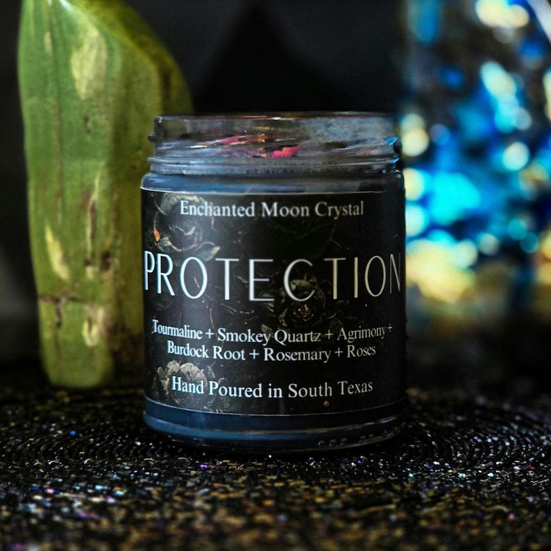 May include: A black candle jar with a white label that reads "Enchanted Moon Crystal PROTECTION Tourmaline + Smokey Quartz + Agrimony Burdock Root + Rosemary + Roses Hand Poured in South Texas".