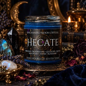 Hand-poured Hecate goddess candle photographed in a mystical gothic setting. The cobalt blue glass candle jar features an elaborate black and antique gold celestial label, surrounded by black crystals, lavender, dried botanicals, glowing candlelight, and a luminous full moon in the background. Inspired by Hecate, the Greek goddess of witchcraft and crossroads, this crystal intention candle evokes moon magic, shadow work, divination, spiritual protection, and transformation