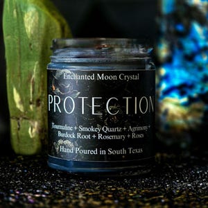 May include: A black candle jar with a white label that reads "Enchanted Moon Crystal PROTECTION Tourmaline + Smokey Quartz + Agrimony + Burdock Root + Rosemary + Roses Hand Poured in South Texas".
