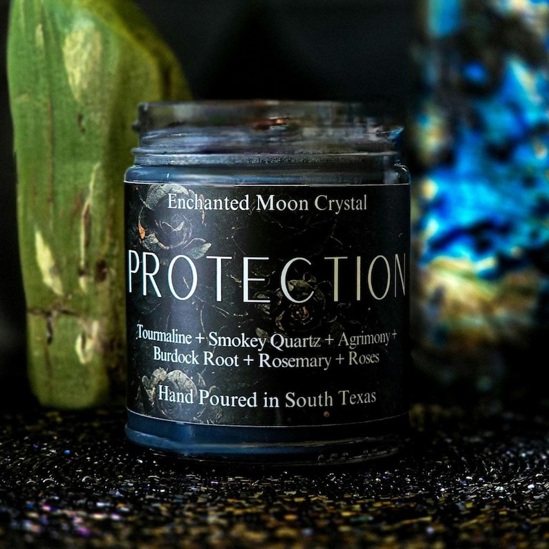 May include: A black candle jar with a white label that reads "Enchanted Moon Crystal PROTECTION Tourmaline + Smokey Quartz + Agrimony + Burdock Root + Rosemary + Roses Hand Poured in South Texas".