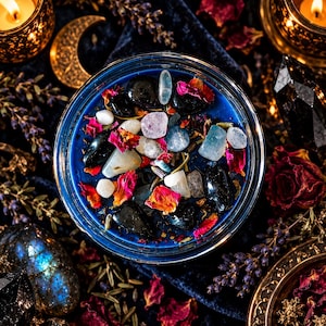 Detailed overhead photograph of the Hecate crystal intention candle with a vibrant royal blue wax surface topped with smooth black obsidian, pale moonstone crystals, colorful rose petals, and botanical herbs. The round glass jar rests in an elaborate gothic altar arrangement featuring a gold crescent moon, deep burgundy roses, dark velvet textures, and candlelit accents. A handmade witchcraft candle celebrating Hecate, the dark feminine, spiritual intuition, protection, and transformative magic.