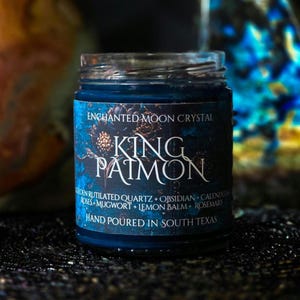 May include: A dark blue glass jar with a label that reads "ENCHANTED MOON CRYSTAL KING PATMON." The label lists ingredients like golden rutilated quartz and rosemary. The text "HAND POURED IN SOUTH TEXAS" is also visible.