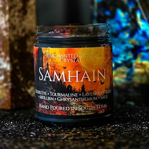May include: A glass jar candle with a black label featuring an orange and black illustration of a graveyard, bats, and a full moon. The text reads Enchanted Moon Crystal, Samhain, Tigers Eye, Tourmaline, Lavender, Roses, Mullien, Chrysanthemum, Sage, and Hand Poured In South Texas.