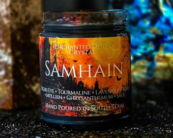 May include: A glass jar candle with a black label featuring an orange and black illustration of a graveyard, bats, and a full moon. The text reads Enchanted Moon Crystal, Samhain, Tigers Eye, Tourmaline, Lavender, Roses, Mullien, Chrysanthemum, Sage, and Hand Poured In South Texas.