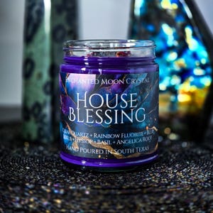 Pu&ograve; includere: Una candela in barattolo di vetro con cera viola scuro. L'etichetta recita "HOUSE BLESSING" con testo aggiuntivo sugli ingredienti e l'origine. La candela ha un design celeste con accenti blu, viola e oro. "HAND POURED IN SOUTH TEXAS" &egrave; stampato sull'etichetta.