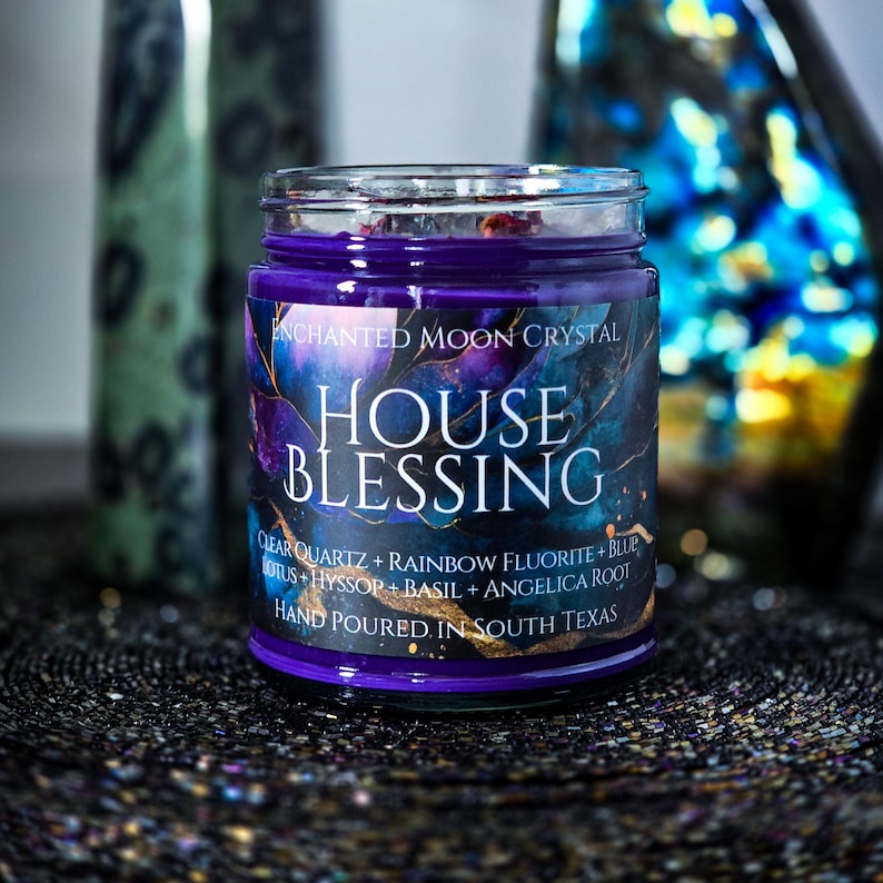 Pu&ograve; includere: Una candela in barattolo di vetro con cera viola scuro. L'etichetta recita "HOUSE BLESSING" con testo aggiuntivo sugli ingredienti e l'origine. La candela ha un design celeste con accenti blu, viola e oro. "HAND POURED IN SOUTH TEXAS" &egrave; stampato sull'etichetta.