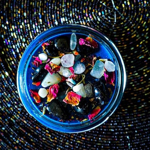 Close top view of the Hecate candle showing vivid blue wax decorated with Moonstone, Lavender, Mugwort, Thyme, and Rosemary. White stones and colorful dried botanicals create a dramatic contrast against the dark blue candle.