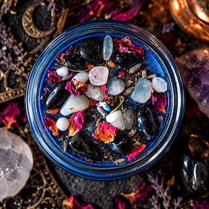 Overhead view of a handcrafted Hecate goddess candle featuring vivid midnight blue wax adorned with polished black obsidian stones, pale moonstone crystals, dried red and pink rose petals, and scattered ritual herbs. Presented in a round glass candle jar surrounded by ornate gold moon decorations, warm candlelight, and dark mystical altar accessories. A crystal-adorned intention candle inspired by Greek goddess Hecate, shadow work, intuition, spiritual protection, and crossroads magic.