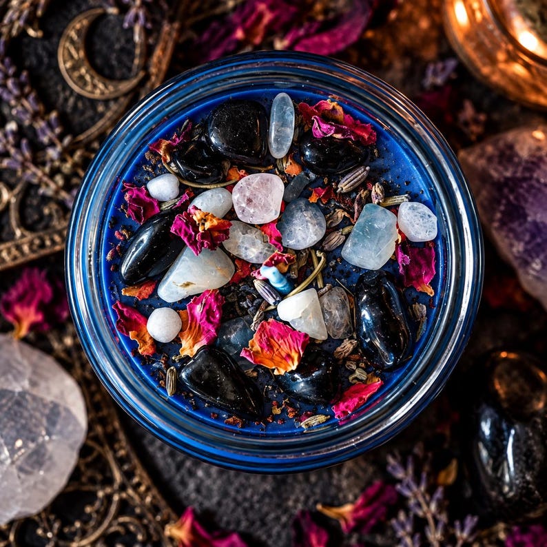 Overhead view of a handcrafted Hecate goddess candle featuring vivid midnight blue wax adorned with polished black obsidian stones, pale moonstone crystals, dried red and pink rose petals, and scattered ritual herbs. Presented in a round glass candle jar surrounded by ornate gold moon decorations, warm candlelight, and dark mystical altar accessories. A crystal-adorned intention candle inspired by Greek goddess Hecate, shadow work, intuition, spiritual protection, and crossroads magic.