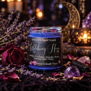 A blue glass candle jar labeled Enchanted Moon Crystal Witching Hour, containing Amethyst, Tourmaline, Lavender, Roses, Rosemary, Basil, and Violets. The label states it is hand poured in South Texas.