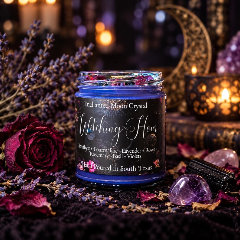 A blue glass candle jar labeled Enchanted Moon Crystal Witching Hour, containing Amethyst, Tourmaline, Lavender, Roses, Rosemary, Basil, and Violets. The label states it is hand poured in South Texas.