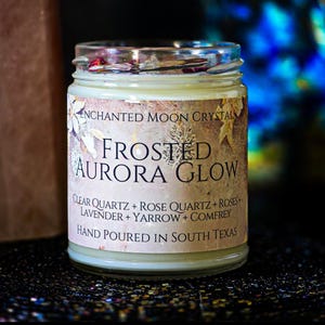 May include: A clear glass candle jar filled with off-white wax and topped with dried flower petals. The label reads Enchanted Moon Crystal, Frosted Aurora Glow, Clear Quartz, Rose Quartz, Roses, Lavender, Yarrow, Comfrey, Hand Poured In South Texas.