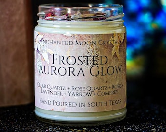 May include: A clear glass candle jar filled with off-white wax and topped with dried flower petals. The label reads Enchanted Moon Crystal, Frosted Aurora Glow, Clear Quartz, Rose Quartz, Roses, Lavender, Yarrow, Comfrey, Hand Poured In South Texas.