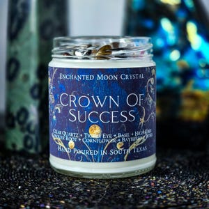 May include: A glass jar candle with a blue and gold label that reads "Crown of Success." The label also says "Enchanted Moon Crystal" and lists ingredients. The candle is hand-poured in South Texas.
