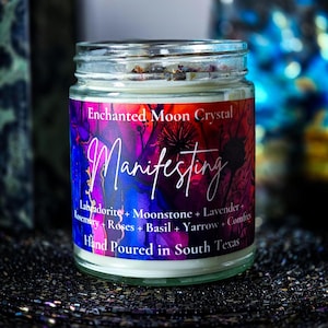 Op de afbeelding: Een glazen potkaars met een kleurrijk etiket. Op het etiket staat "Enchanted Moon Crystal Manifesting" met ingredi&euml;ntnamen. De kaars is met de hand gegoten in South Texas. De kaars heeft een witte was en is bedekt met gedroogde botanische producten.
