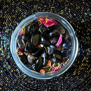 May include: A clear glass jar filled with black stones, dried rose petals, and herbs. The jar is sitting on a black and silver beaded surface.