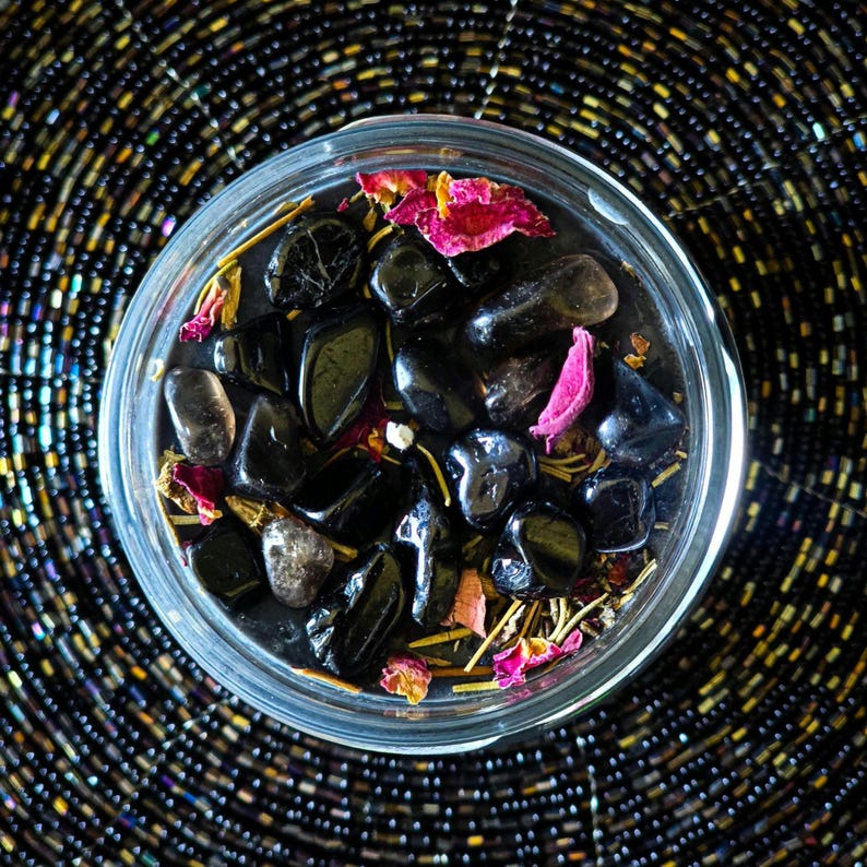 May include: A clear glass jar filled with black stones, dried rose petals, and herbs. The jar is sitting on a black and silver beaded surface.