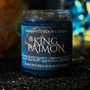 May include: A blue glass jar with a label that reads "ENCHANTED MOON CRYSTAL" and "KING PAIMON" in white text. The label also lists ingredients like golden rutilated quartz and rosemary. The jar is hand-poured in South Texas.