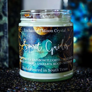 May include: A glass jar candle with a label that reads "Enchanted Moon Crystal Spirit Guides." The label features a cosmic design with teal and gold accents. The candle is hand-poured in South Texas and contains quartz, rainbow fluorite, and blue lotus.
