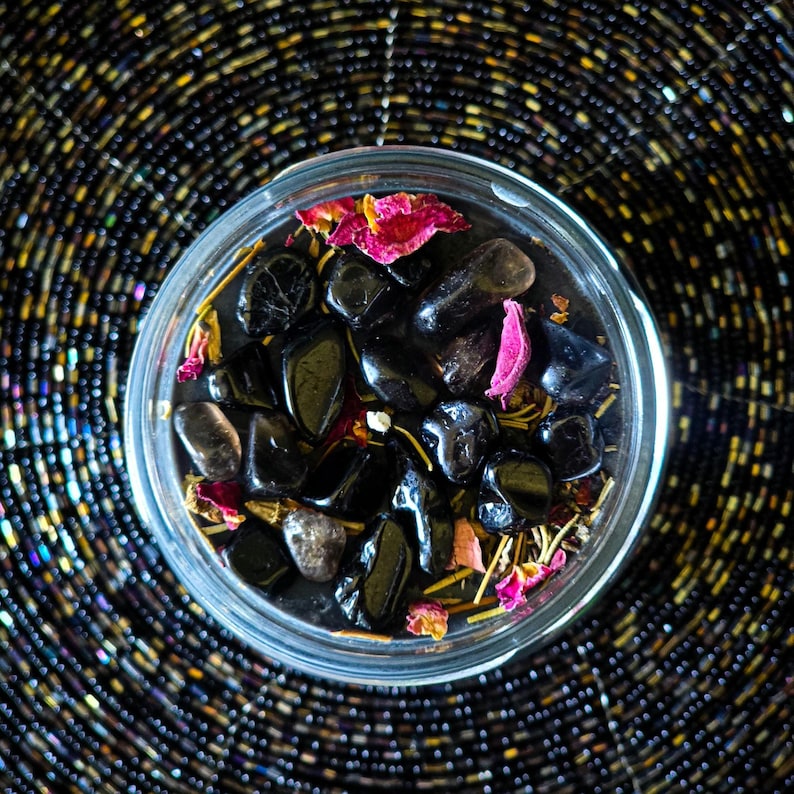 May include: A glass jar filled with black stones, dried rose petals, and other natural elements. The jar is sitting on a black and gold patterned surface.